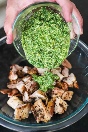 A person transferring homemade pesto sauce from a small glass bowl into a larger glass bowl filled with grilled chicken thighs that have been cut into bite-size pieces.