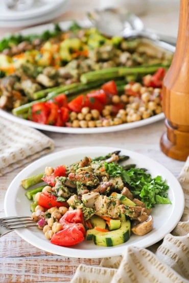 A close-up view of a grilled chicken salad that also has cucumbers, tomato wedges, mushrooms, and asparagus on the plate.