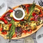 An overhead view of a wooden oval platter filled with grilled vegetables that have been drizzled with Italian dressing in a white bowl with a spoon on it.