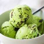 A straight-on view of a white ice cream bowl filled with four scoops of homemade mint chocolate chip ice cream with a spoon inserted into the back of the bowl.