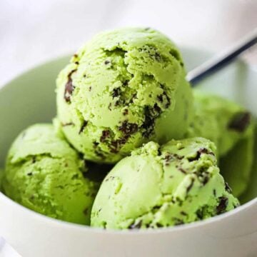 A straight-on view of a white ice cream bowl filled with four scoops of homemade mint chocolate chip ice cream with a spoon inserted into the back of the bowl.