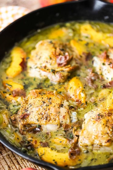 A close-up view of a large black cast-iron skillet that is filled with braised chicken and peaches with crispy prosciutto on top.