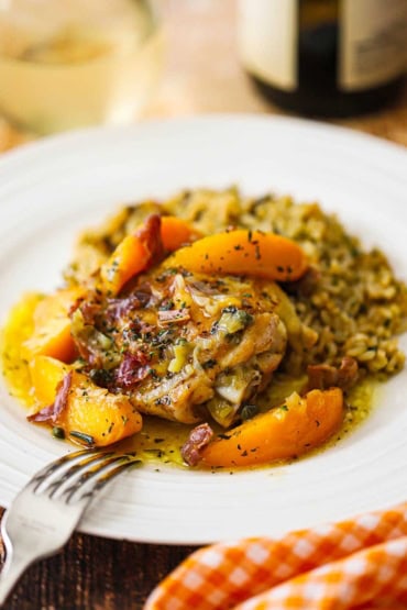 A close-up view a white dinner plate filled with a serving of braised chicken thighs topped with savory peaches served next to wild rice.