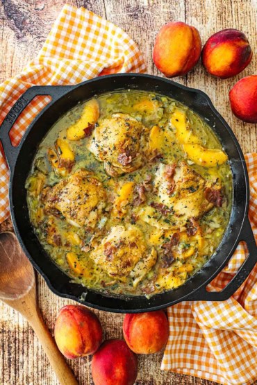 An overhead view of large black cast-iron skillet that is filled with braised chicken and peaches with a wooden spoon and fresh peaches nearby.
