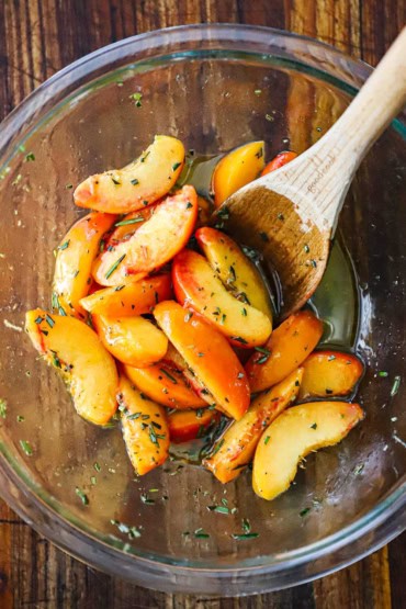 An overhead view of a glass bowl that is filled with slices of fresh peaches that have been marinated in sherry vinegar, olive oil, and fresh herbs.