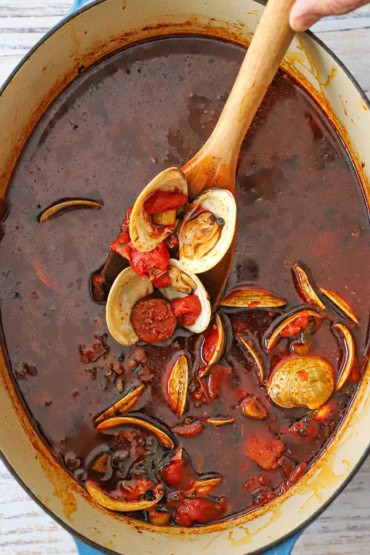 An overhead view of a tomato-based clam chowder in a large oval Dutch oven with a wooden spoon being used to raise up a helping of the chowder.