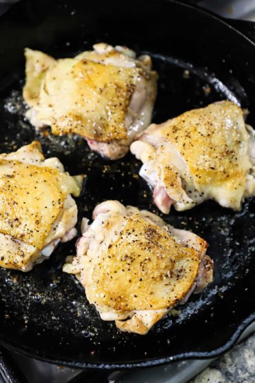 A cast-iron skillet filled with four skin-on chicken thighs that are being seared in oil and the skin on top is golden and crispy.
