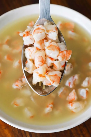 A overhead view of a large silver slotted spoon holding up pieces of shrimp that have been cooked in freshly squeezed lime juice that appear below the raised spoon.