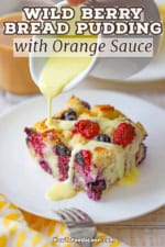 A person pouring a thick orange sauce over the top of a sliced of wild berry bread pudding that is sitting on a white dessert plate.