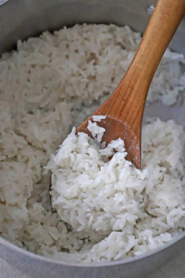 A close-up view of a silver saucepan that is filled with easy coconut rice that has been cooked and stirred with a wooden spoon.