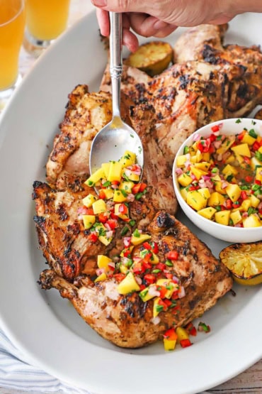 A person using a large silver serving spoon to scatter homemade mango salsa over the tops of whole jerk chicken legs on a large white platter.
