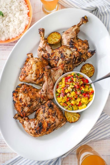 An overhead view of a large white platter that is filled with a row of lightly charred Jamaican jerk chicken with a bowl of mango salsa next to the whole chicken legs.