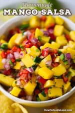 A close-up view of mango salsa that is in a white bowl with tortilla chips and lime wedges surrounding it.