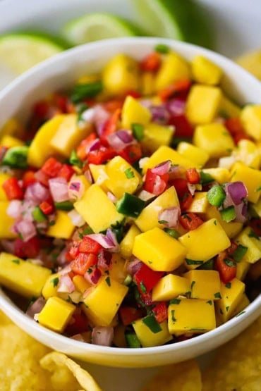 A close-up view of mango salsa that is in a white bowl with tortilla chips and lime wedges surrounding it.
