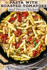 An overhead view of a large black cast-iron skillet that is filled with pasta with roasted tomatoes and pesto chicken.