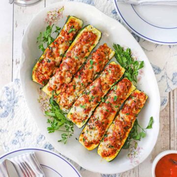 An overhead view of an antique platter filled with stuffed zucchini boats that are topped with melty cheese and surrounded by sprigs of parsley.