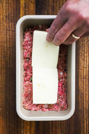 A person layering two thick slices of Fontina cheese into a loaf pan that the bottom has been packed with an uncooked meatloaf mixture.