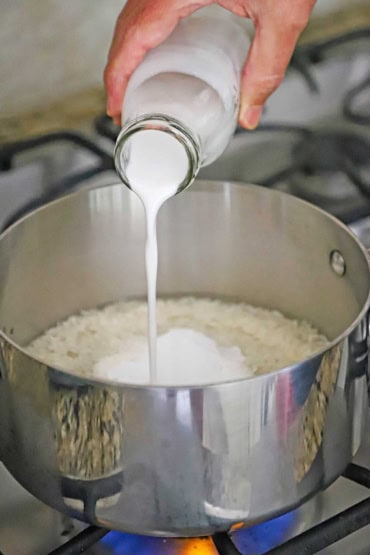 A person pouring unsweetened coconut milk from a small glass milk jug into a medium-sized silver saucepan that is filled with uncooked Jasmine rice.