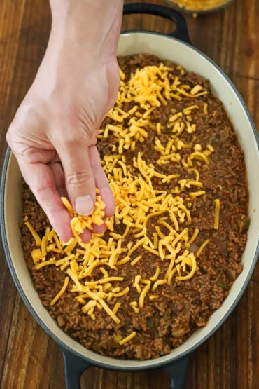 An overhead view of a person sprinkling shredded cheddar cheese over the top of an oval casserole dish filled with a sloppy Joe mixture.