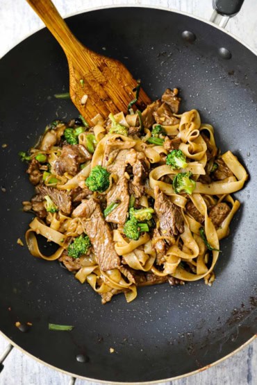 An overhead view of a non-stick wok that is filled with a stir-fried beef and broccoli chow fun intermingled with cooked wide rice noodles.