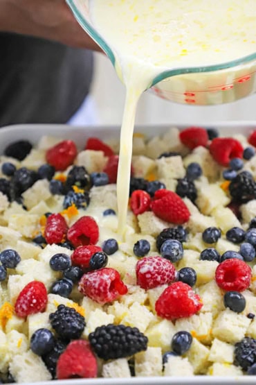 A person pouring an egg and cream mixture from a large glass measuring cup into a square baking dish that is filled with cubed bread and mixed berries.