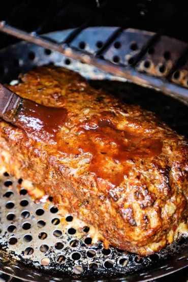 A close-up view of a cooked cheese-stuffed smoked meatloaf in a grill pan inside the smoker and a person is basting the top of the loaf with a balsamic glaze.
