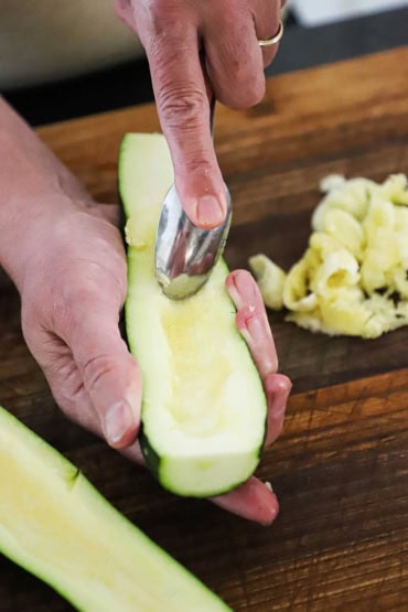 A person holding a zucchini that has been cut in half lengthwise and is using a spoon to scrape out the seeds.