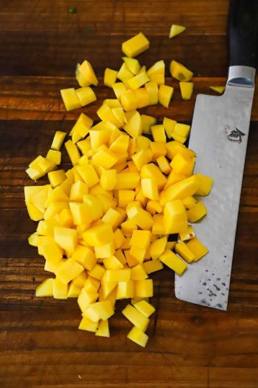 An overhead view of fresh mango that has been cut into bite-sized pieces on a cutting board with a large Japanese-style knife next to the cut fruit.