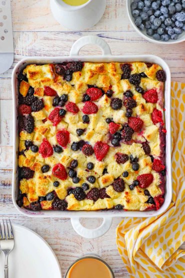 An overhead view of a square white baking dish that is filled with a fully cooked wild berry bread pudding and is surrounded by a bowl of blueberries and a festive linen.