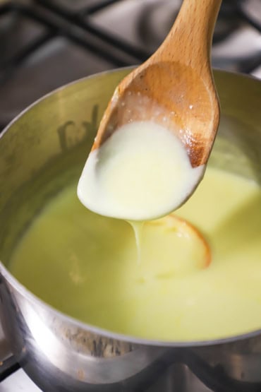 A close-up view of a wooden spoon that is being lifted from a saucepan that is filled with an orange sauce and that same sauce is dripping from the spoon.