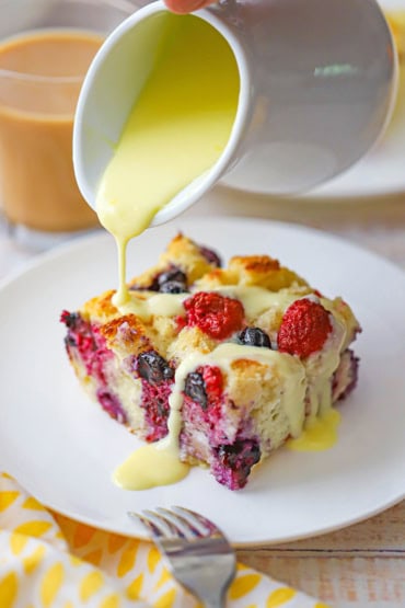 A person pouring a thick orange sauce over the top of a sliced of wild berry bread pudding that is sitting on a white dessert plate.