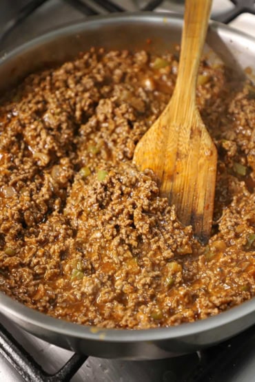A straight-on view of a large silver skillet filled with a sloppy Joe mixture and a wooden spatula in the middle of it.