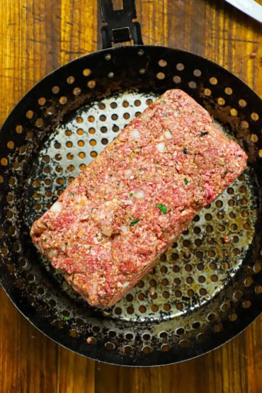 An overhead view of a rectangular loaf of an uncooked meatloaf resting in a circular grill pan that has holes throughout it.