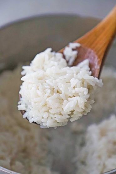 A close-up view of a wooden spoon that is holding a serving of easy coconut rice over a saucepan that is filled with the same rice.
