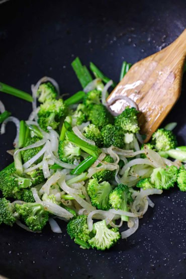 A close-up view of strips of white onion, pieces of green onion, and broccoli florets that are being stir-fried in a wok with a wooden spatula.
