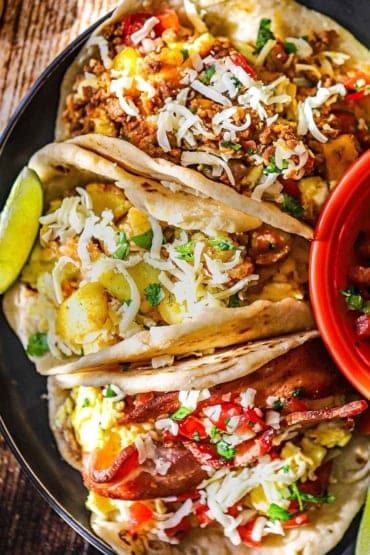 An overhead view of a black plate that is filled with three types of stuffed breakfast tacos that are topped with shredded Monterey Jack cheese.