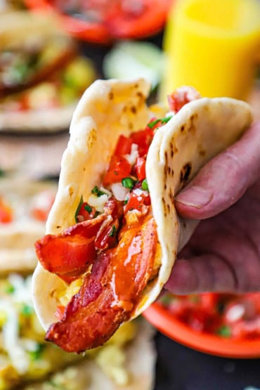 A person holding up a breakfast taco that is made with a homemade flour tortilla and is stuffed with eggs, bacon, cheese, and topped with pico de gallo.