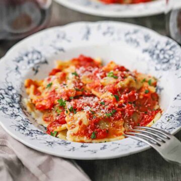A close-up view of an antique bowl that is filled with a serving of homemade beef ravioli that is topped with a tomato sauce, shredded parmesan cheese, and chopped basil.