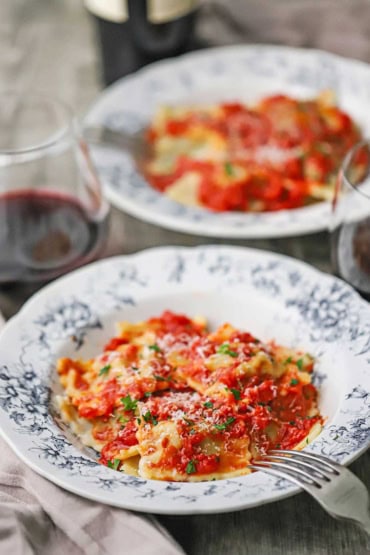 A straight-on view of two antique bowls that are each filled with a serving of homemade beef ravioli with tomato sauce and grated parmesan cheese.