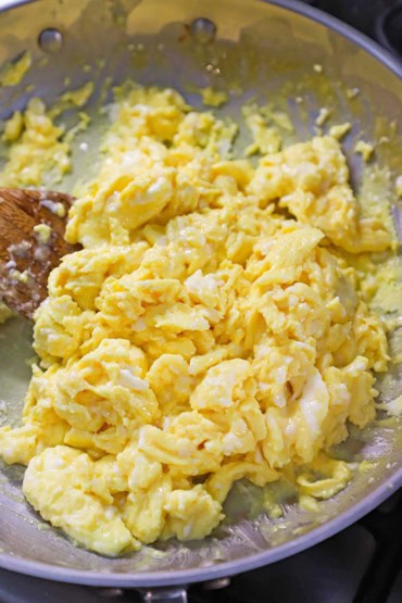 A close-up view of creamy and fluffy scrambled eggs in a stainless steel skillet with a wooden spatula off to the side.