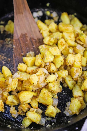 A close-up view of sautéed cubed yellow potatoes and onion in a cast-iron skillet with a wooden spatula off to the side.