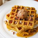 A straight-on view of a stack of three pumpkin waffles that are topped with a dollop of whipped cinnamon honey butter, maple syrup, and chopped pecans.