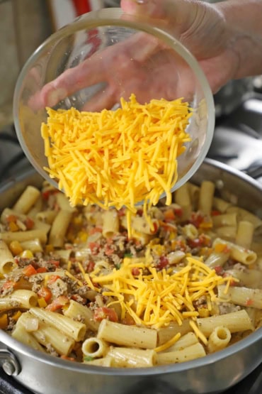 A person transferring shredded cheddar cheese from a glass bowl into a skillet filled with cooked rigatoni in a simmering Tex-Mex beef sauce.