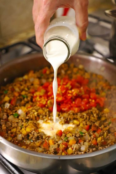 A person pouring half and half from a small milk bottle into a skillet filled with a Tex-Mex beef mixture with chopped pepper, tomatoes, and broth.