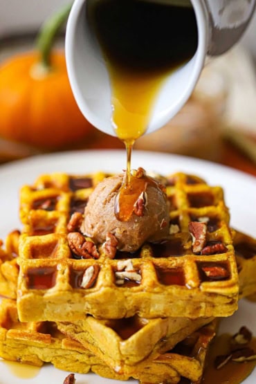 A person pouring pure maple syrup from a small white pitcher over a pile of pumpkin waffles with whipped cinnamon honey waffles.