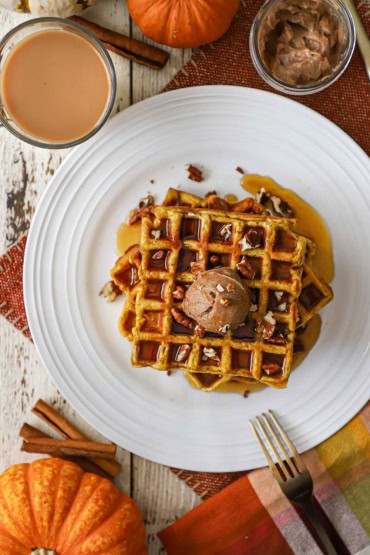 An overhead view of a white plate that is filled with a stack of pumpkin waffles with whipped cinnamon honey butter and a small pumpkin and cup of coffee nearby.