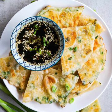 An overhead view of quarted scallion pancakes on a plate with a bowl filled with a dipping sauce.