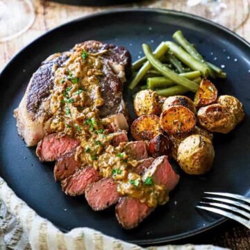 A straight-on view of a reverse-sear ribeye steak with gorgonzola on a black dinner plate alongside a helping of roasted potatoes and steamed green beans.