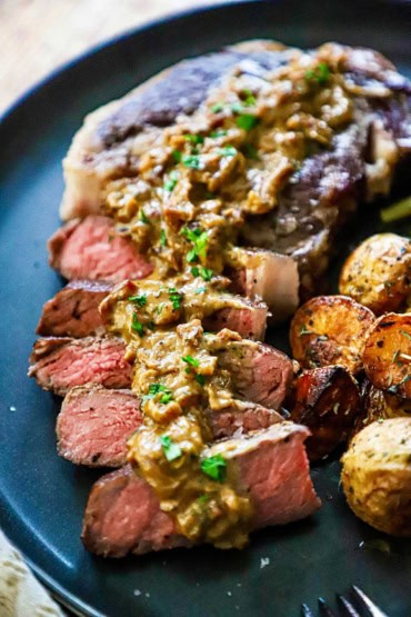 A straight-on view of reverse-seared ribeye with gorgonzola sauce on a black dinner plate alongside a helping of roasted potatoes.