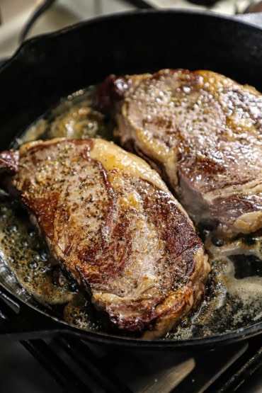 A close-up view of two large ribeye steaks that are being seared in a hot cast-iron skillet with sizzling butter and oil under the meat.